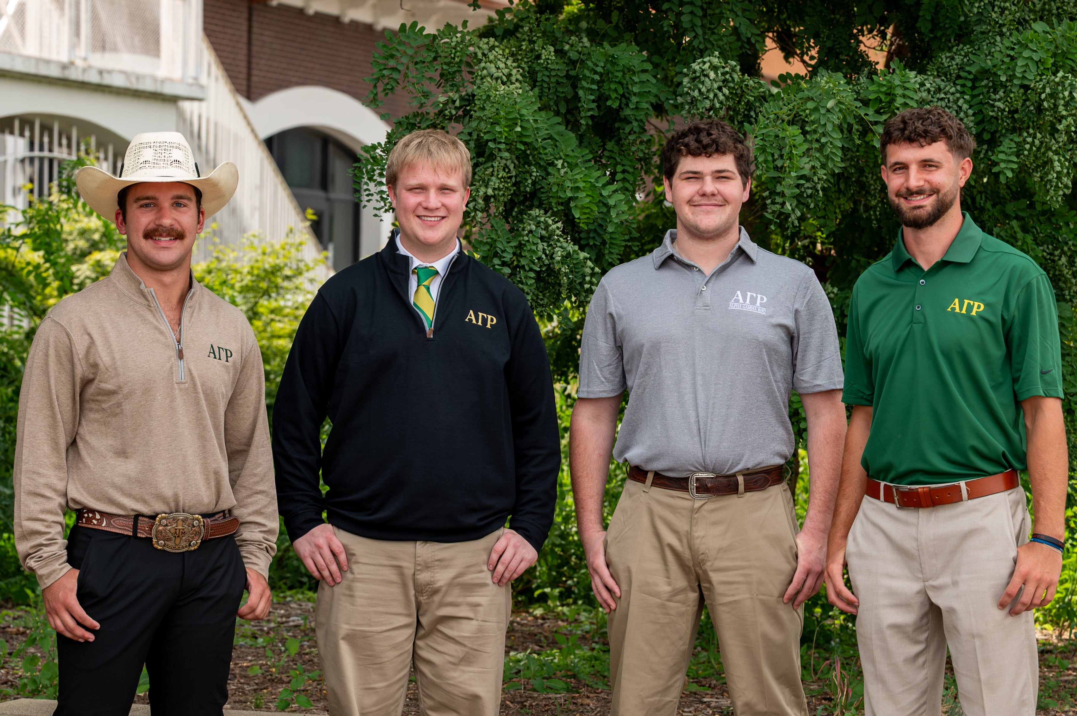 Four men standing outdoors in front of a building and greenery, wearing matching branded shirts in beige, navy, light gray, and green, with khaki pants and brown shoes.